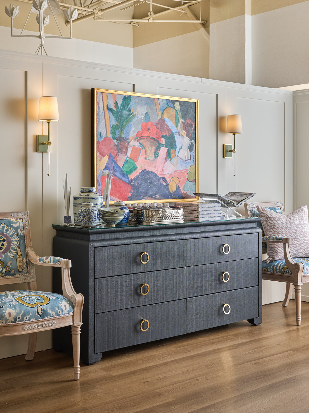 A coordinated showroom vignette with artwork, brass sconces, a dark chest and patterned accent chairs.